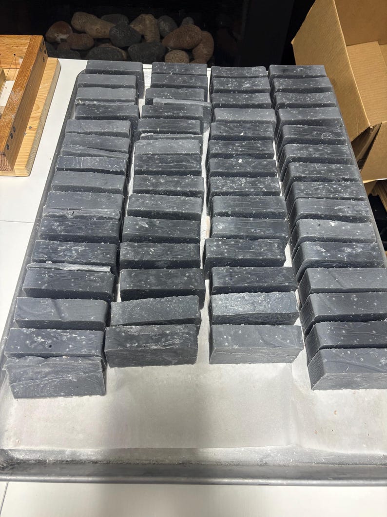 May include: A large collection of rectangular charcoal-colored soap bars arranged in neat rows on a metal tray. Each bar features a solid dark gray base with subtle white speckles distributed across the surface, creating a uniform appearance.