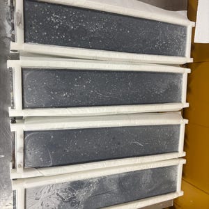 May include: Four rectangular soap molds arranged in a vertical stack. Each mold features a white plastic frame containing a dark gray, solid block of soap with visible white speckles and swirling patterns on the surface, held by metal hardware.