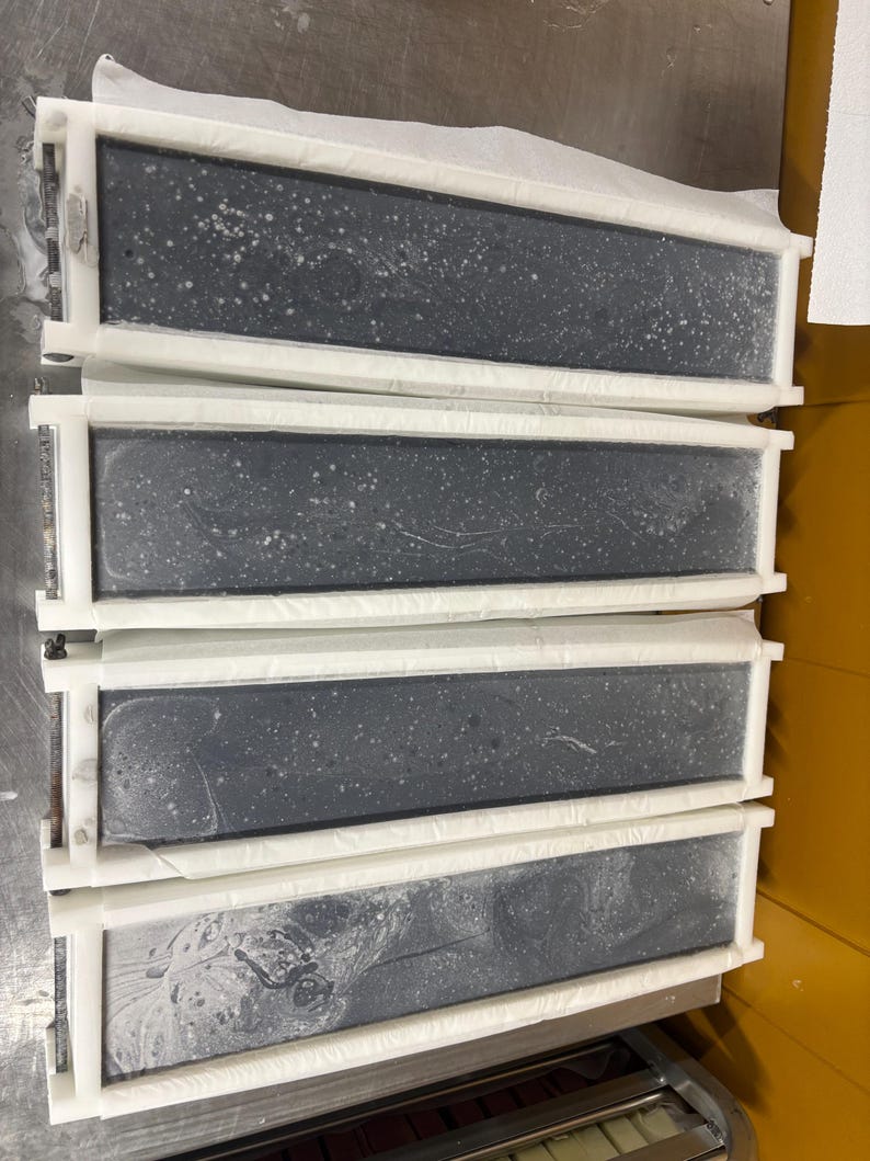 May include: Four rectangular soap molds arranged in a vertical stack. Each mold features a white plastic frame containing a dark gray, solid block of soap with visible white speckles and swirling patterns on the surface, held by metal hardware.