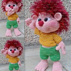 Könnte beinhalten: Eine gehäkelte Puppe mit rosa Haut, roten Haaren, einem gelben Shirt und einer grünen Hose. Die Puppe steht auf einer grauen Oberfläche.
