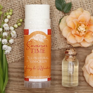 Op de afbeelding: Een stick met volledig natuurlijke zonnebrandcrème met het opschrift "Summer Time" en de woorden "Juicy & Refreshing". De afbeelding bevat perzikrozen, een kleine glazen fles en een takje witte bloemen.