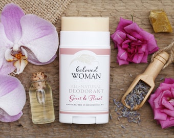 Könnte beinhalten: Ein weißer Deodorant-Stick mit dem Text "beloved WOMAN" und "Sweet & Floral" ist von lila Orchideen, rosa Rosen, einer kleinen Glasflasche und einer Holzschaufel mit Lavendelknospen umgeben. Der Hintergrund ist eine Holzoberfläche.