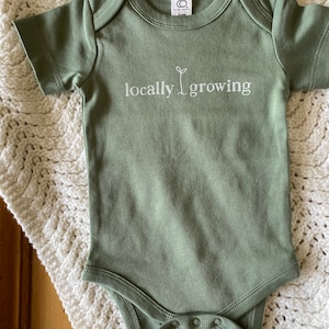 May include: Sage green baby onesie with the words "locally growing" printed in white. The onesie has short sleeves and snaps at the bottom. The onesie is made of soft material and is laying on a white crocheted blanket.