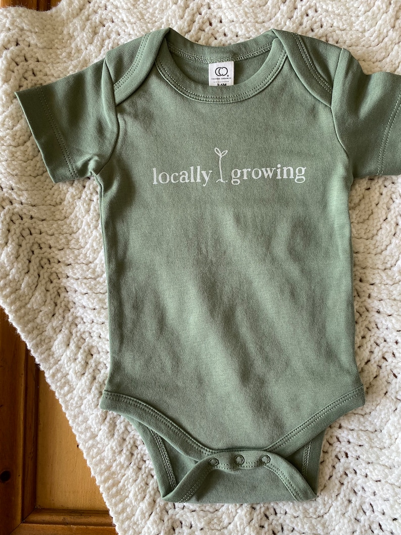 May include: Sage green baby onesie with the words "locally growing" printed in white. The onesie has short sleeves and snaps at the bottom. The onesie is made of soft material and is laying on a white crocheted blanket.