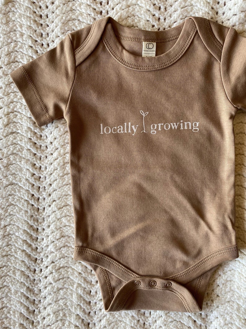 May include: A short-sleeved, brown baby bodysuit with the words "locally growing" in white script. The bodysuit has a round neckline and snap closures at the bottom. The garment is displayed on a white, textured surface.