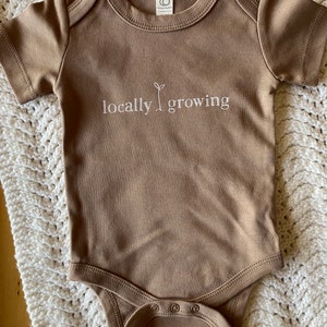 May include: A brown baby onesie with short sleeves and snap closures. The text "locally growing" is printed in white on the front, with a small sprout graphic. The onesie is made of soft material.