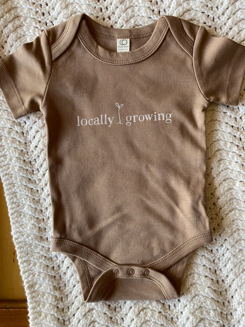 May include: A brown baby onesie with short sleeves and snap closures. The text "locally growing" is printed in white on the front, with a small sprout graphic. The onesie is made of soft material.