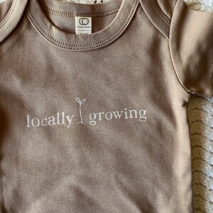 May include: A tan baby onesie with short sleeves. The words "locally growing" are embroidered in white, with a small sprout graphic. The onesie has a round neckline and a tag that reads "Organic Children".
