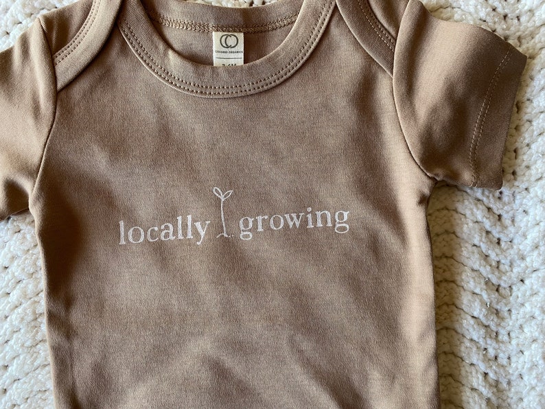 May include: A tan baby onesie with short sleeves. The words "locally growing" are embroidered in white, with a small sprout graphic. The onesie has a round neckline and a tag that reads "Organic Children".