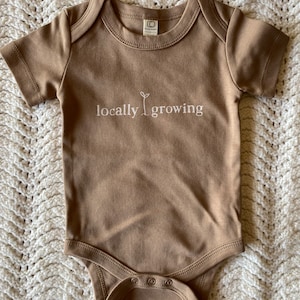 May include: A tan-colored baby onesie with short sleeves and snap closures. The text "locally growing" is printed in white, with a small plant graphic. The onesie is made of soft material.