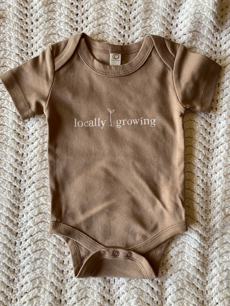 May include: A tan-colored baby onesie with short sleeves and snap closures. The text "locally growing" is printed in white, with a small plant graphic. The onesie is made of soft material.