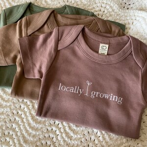 May include: Three short-sleeved baby onesies in shades of green, brown, and mauve. The front onesie features the text "locally growing" with a sprout graphic. The onesies are laid on a white, crocheted blanket.