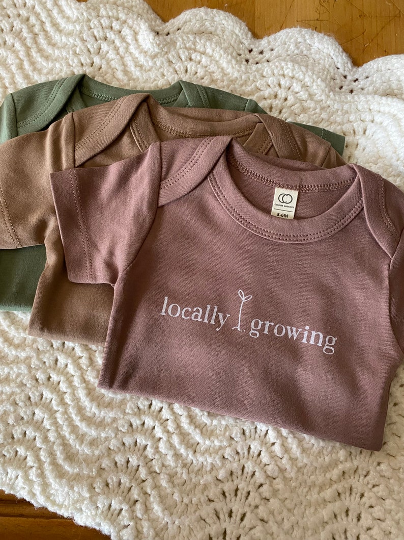 May include: Three short-sleeved baby onesies in shades of green, brown, and mauve. The front onesie features the text "locally growing" with a sprout graphic. The onesies are laid on a white, crocheted blanket.