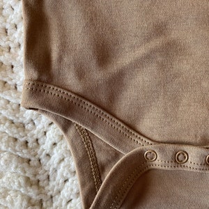 May include: Close-up of a tan-colored bodysuit. The fabric has a ribbed texture, with visible stitching along the edges and snap closures at the bottom. The bodysuit is likely for a baby or toddler.