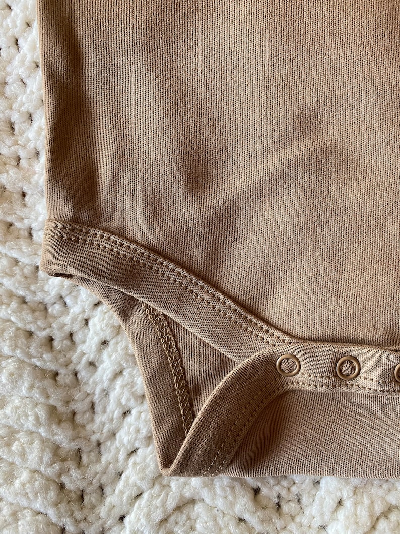 May include: Close-up of a tan-colored bodysuit. The fabric has a ribbed texture, with visible stitching along the edges and snap closures at the bottom. The bodysuit is likely for a baby or toddler.