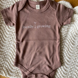 May include: A mauve-colored baby onesie with short sleeves and a round neckline. The text "locally growing" is embroidered in white across the chest, with a small plant graphic. The onesie has snap closures at the bottom and a tag at the back of the neck.