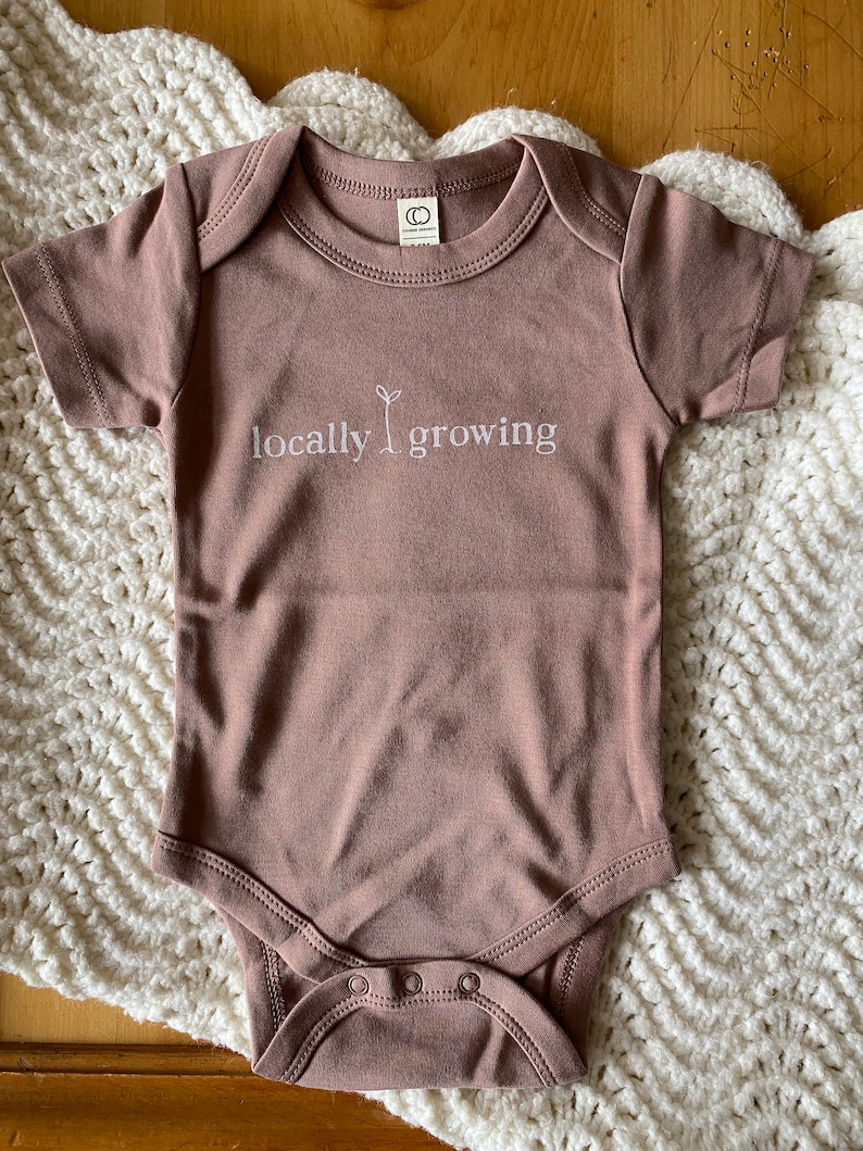 May include: A mauve-colored baby onesie with short sleeves and a round neckline. The text "locally growing" is embroidered in white across the chest, with a small plant graphic. The onesie has snap closures at the bottom and a tag at the back of the neck.