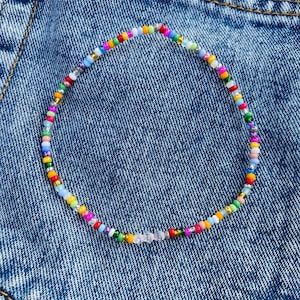May include: A colorful beaded necklace with a variety of small, round beads in shades of red, yellow, green, blue, and purple. The necklace is displayed on a denim background.