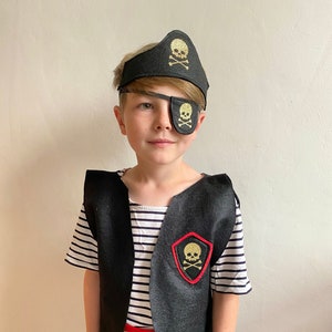 Peut inclure: Un enfant déguisé en pirate portant un gilet noir avec un écusson de crâne et d'os croisés dorés, un cache-œil noir avec un crâne doré, un chapeau de pirate noir avec un crâne et des os croisés dorés, une ceinture rouge et une chemise rayée.
