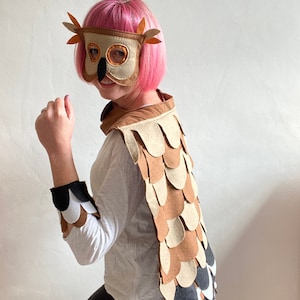May include: A child wearing a brown and beige owl costume with a felt owl mask. The costume features a brown and beige feathered cape with a black and white trim. The mask has large brown eyes and an orange beak.