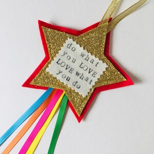 May include: A star-shaped ornament with a red felt base, a gold glitter star in the center, and a white fabric square with the text "do what you LOVE love what you do." Rainbow ribbons and a gold ribbon are attached.