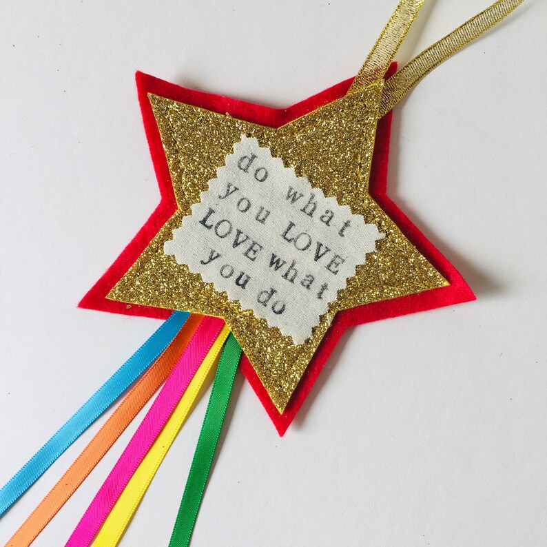 May include: A star-shaped ornament with a red felt base, a gold glitter star in the center, and a white fabric square with the text "do what you LOVE love what you do." Rainbow ribbons and a gold ribbon are attached.