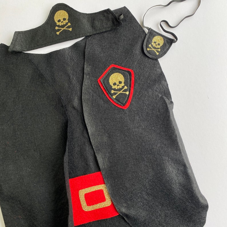 K&ouml;nnte beinhalten: Schwarzes Filz-Piratenkost&uuml;m mit einem roten und goldenen Totenkopf- und Knochen-Design. Das Kost&uuml;m beinhaltet einen Umhang, einen Hut und ein Schwert.