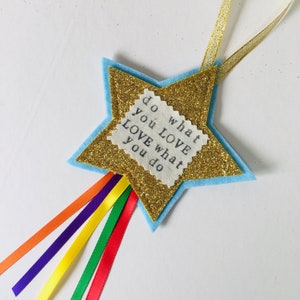 May include: A star-shaped ornament with a gold glitter center and a light blue felt border. The ornament has a small rectangular patch with the text "do what you LOVE LOVE what you do". Rainbow ribbons and a gold ribbon hang from the star.