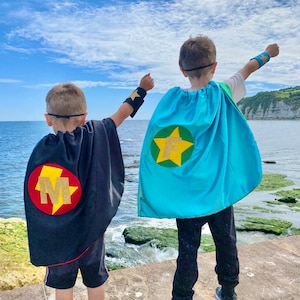 May include: Two children wearing superhero capes with personalised letters. The boy on the left is wearing a black cape with a red circle and the letter "M" in gold. The boy on the right is wearing a blue cape with a green circle and the letter "F" in gold.