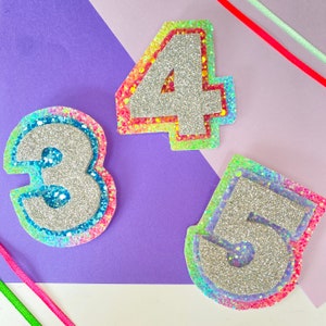 May include: Three sparkly silver numbers, 3, 4, and 5, with rainbow glitter borders. The numbers are on a purple background.
