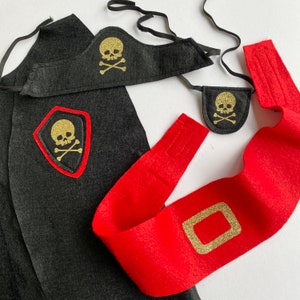 K&ouml;nnte beinhalten: Ein Piratenkost&uuml;m-Set aus schwarzem Filz mit Totenkopf- und Knochenmotiv. Das Set beinhaltet eine Kappe, eine Augenklappe und eine Sch&auml;rpe.