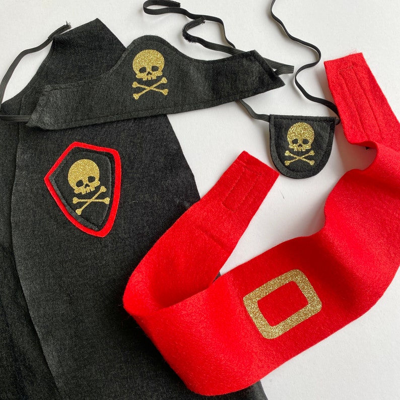 K&ouml;nnte beinhalten: Ein Piratenkost&uuml;m-Set aus schwarzem Filz mit Totenkopf- und Knochenmotiv. Das Set beinhaltet eine Kappe, eine Augenklappe und eine Sch&auml;rpe.