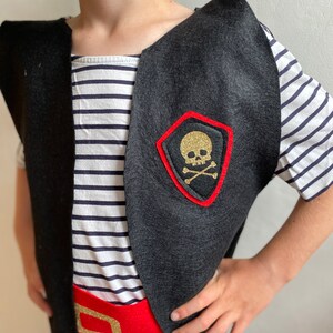 K&ouml;nnte beinhalten: Eine schwarze Filz-Piratenweste mit einem roten Filzg&uuml;rtel und einem goldenen Totenkopf- und Knochen-Aufn&auml;her. Die Weste hat einen weiten Ausschnitt und wird &uuml;ber einem gestreiften Hemd getragen.