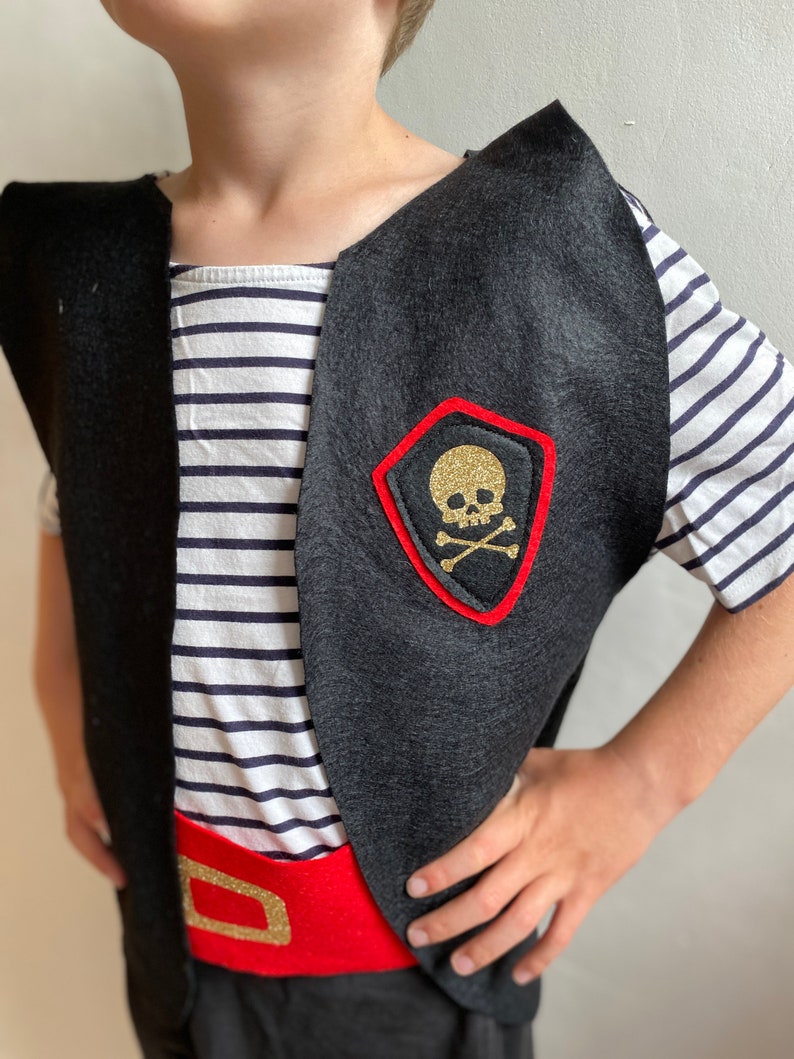 K&ouml;nnte beinhalten: Eine schwarze Filz-Piratenweste mit einem roten Filzg&uuml;rtel und einem goldenen Totenkopf- und Knochen-Aufn&auml;her. Die Weste hat einen weiten Ausschnitt und wird &uuml;ber einem gestreiften Hemd getragen.