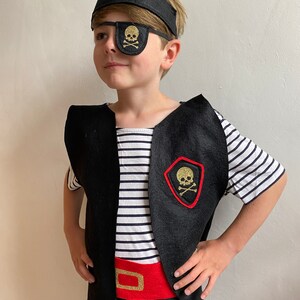 K&ouml;nnte beinhalten: Ein Kind verkleidet als Pirat, das eine schwarze Filz-Weste mit einem goldenen Totenkopf- und Knochen-Aufn&auml;her, eine schwarze Filz-Augenklappe mit einem goldenen Totenkopf- und Knochen-Aufn&auml;her, einen schwarzen Filz-Piratenhut mit einem goldenen Totenkopf- und Knochen-Aufn&auml;her und einen roten Filzg&uuml;rtel mit einer goldenen Schnalle tr&auml;gt.