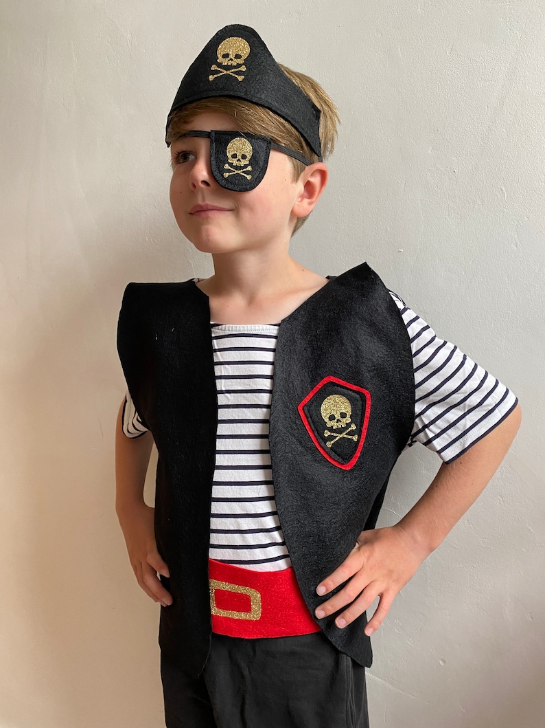 K&ouml;nnte beinhalten: Ein Kind verkleidet als Pirat, das eine schwarze Filz-Weste mit einem goldenen Totenkopf- und Knochen-Aufn&auml;her, eine schwarze Filz-Augenklappe mit einem goldenen Totenkopf- und Knochen-Aufn&auml;her, einen schwarzen Filz-Piratenhut mit einem goldenen Totenkopf- und Knochen-Aufn&auml;her und einen roten Filzg&uuml;rtel mit einer goldenen Schnalle tr&auml;gt.