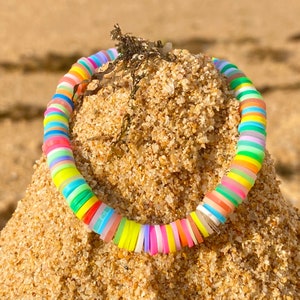 May include: A colorful bracelet made of round, flat, plastic beads. The beads are arranged in a rainbow pattern, with each bead a different color. The bracelet is lying on a pile of sand.