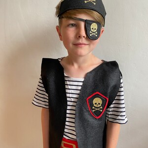 K&ouml;nnte beinhalten: Ein Kind verkleidet als Pirat mit einem schwarzen und goldenen Augenklappen mit Totenkopf und Knochen, einer schwarzen Filz-Piratenm&uuml;tze mit goldenem Totenkopf und Knochen, einer schwarzen Weste mit einem roten und goldenen Totenkopf und Knochen-Aufn&auml;her und einem roten G&uuml;rtel.