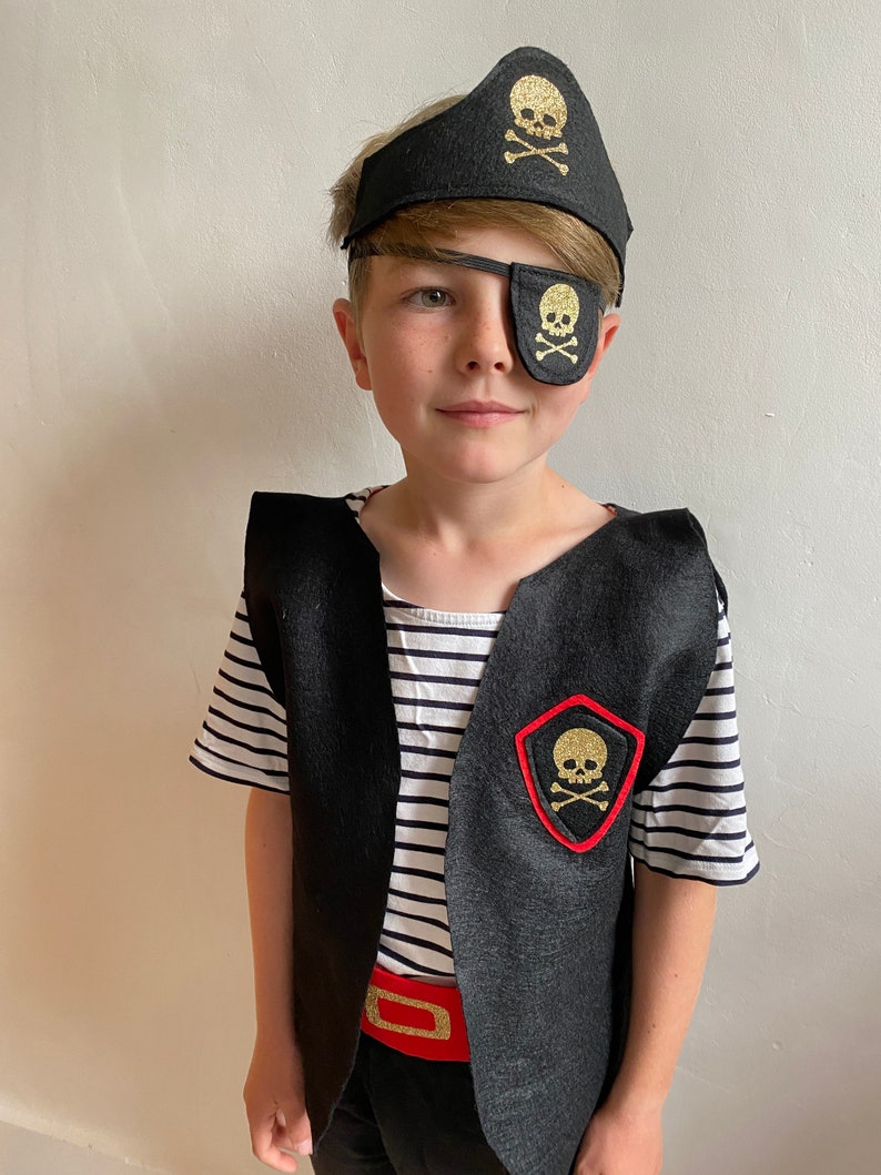 K&ouml;nnte beinhalten: Ein Kind verkleidet als Pirat mit einem schwarzen und goldenen Augenklappen mit Totenkopf und Knochen, einer schwarzen Filz-Piratenm&uuml;tze mit goldenem Totenkopf und Knochen, einer schwarzen Weste mit einem roten und goldenen Totenkopf und Knochen-Aufn&auml;her und einem roten G&uuml;rtel.