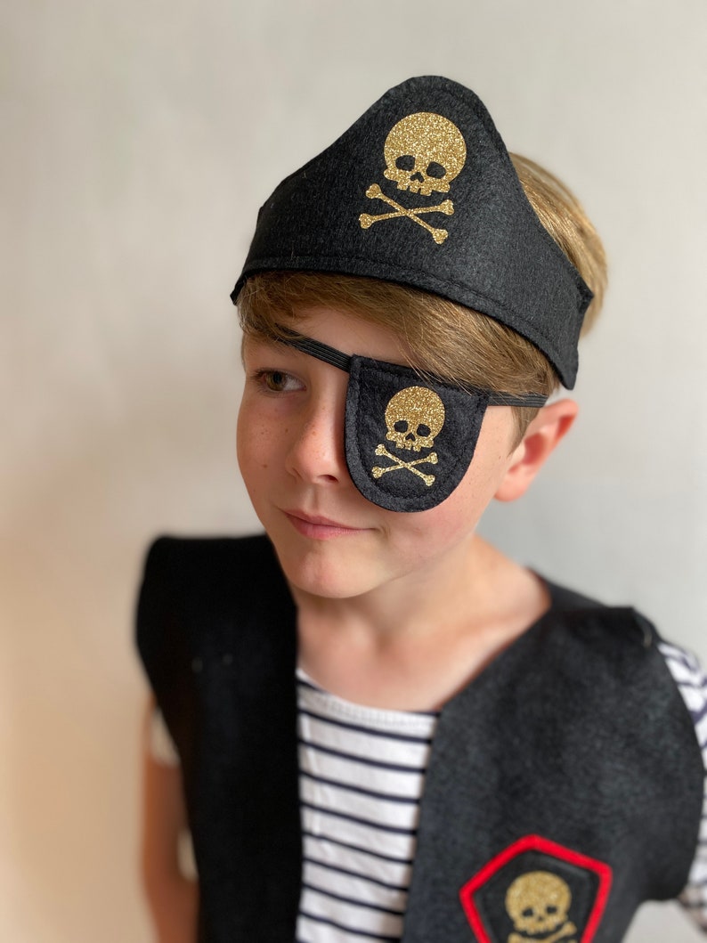K&ouml;nnte beinhalten: Ein Kind tr&auml;gt ein schwarzes Piratenkost&uuml;m aus Filz mit einem goldenen Augenklappen und einem Stirnband mit Totenkopf und Knochen.