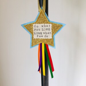 May include: A star-shaped decoration with gold glitter, light blue trim, and the text "do what you LOVE LOVE what you do." It has colorful ribbons in orange, purple, yellow, green, and red. A gold ribbon is at the top for hanging.