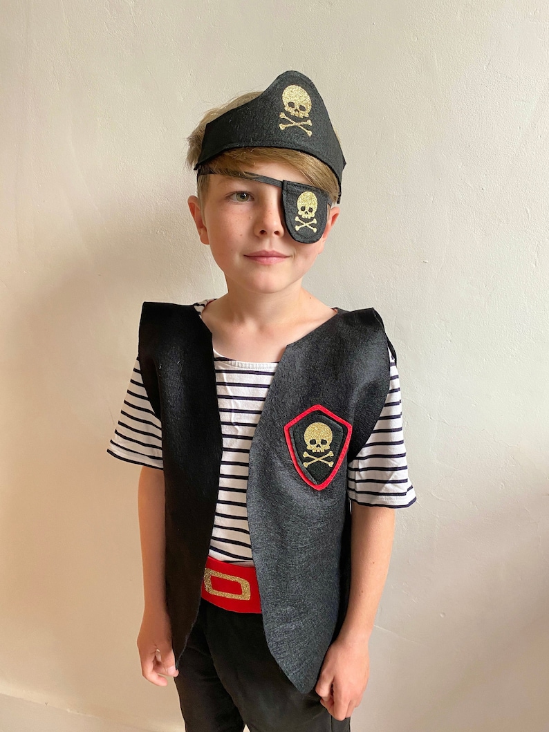 K&ouml;nnte beinhalten: Ein Kind verkleidet als Pirat, das eine schwarze Weste mit einem goldenen Totenkopf- und Knochen-Aufn&auml;her, eine schwarze Augenklappe mit einem goldenen Totenkopf, einen schwarzen Piratenhut mit einem goldenen Totenkopf- und Knochen-Aufn&auml;her, einen roten G&uuml;rtel und ein gestreiftes Hemd tr&auml;gt.
