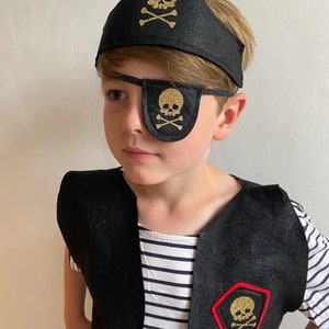 K&ouml;nnte beinhalten: Ein Kind tr&auml;gt ein schwarzes Piratenkost&uuml;m mit einem goldenen Totenkopf-Augenklappen, einem Stirnband und einer Weste.