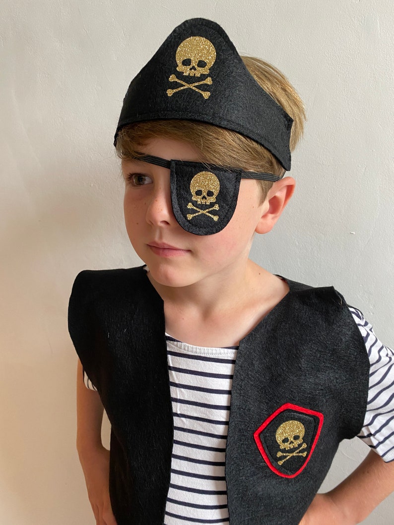 K&ouml;nnte beinhalten: Ein Kind tr&auml;gt ein schwarzes Piratenkost&uuml;m mit einem goldenen Totenkopf-Augenklappen, einem Stirnband und einer Weste.