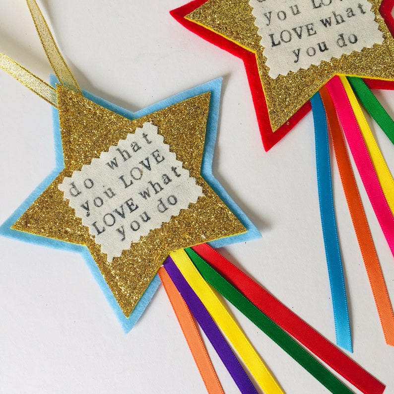 May include: Two star-shaped ornaments with glitter and ribbon. One star is gold glitter on a blue star, the other is gold glitter on a red star. Each has a fabric patch with the text "do what you LOVE LOVE what you do". Rainbow ribbons hang from the stars.