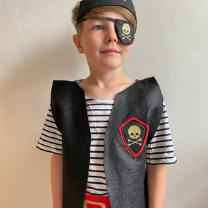 K&ouml;nnte beinhalten: Ein Kind verkleidet als Pirat, das eine schwarze Weste mit einem roten G&uuml;rtel, ein schwarz-wei&szlig; gestreiftes Hemd, eine schwarze Augenklappe mit einem goldenen Totenkopf und gekreuzten Knochen und einen schwarzen Piratenhut mit einem goldenen Totenkopf und gekreuzten Knochen tr&auml;gt.