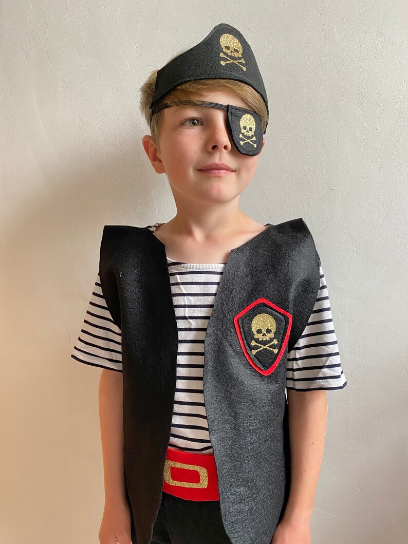 K&ouml;nnte beinhalten: Ein Kind verkleidet als Pirat, das eine schwarze Weste mit einem roten G&uuml;rtel, ein schwarz-wei&szlig; gestreiftes Hemd, eine schwarze Augenklappe mit einem goldenen Totenkopf und gekreuzten Knochen und einen schwarzen Piratenhut mit einem goldenen Totenkopf und gekreuzten Knochen tr&auml;gt.
