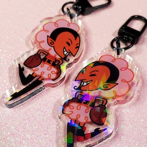 Puede incluir: Dos llaveros iridiscentes de color rosa y rojo con personajes de dibujos animados con cuernos y alas. Los personajes llevan una bolsa de rayas rojas y blancas.