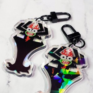 Puede incluir: Dos llaveros de personajes de anime negro y rojo con detalles holográficos iridiscentes. El personaje es un demonio con cuernos y un atuendo rojo y negro. Los llaveros están unidos a llaveros de metal negro.