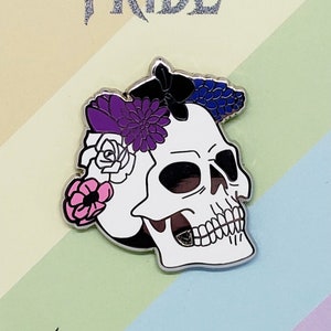 Puede incluir: Una calavera blanca con una corona floral en tonos de morado, rosa y blanco. La calavera está sobre un fondo arcoíris con el texto "Spooky Pride" y "artbykino.com".
